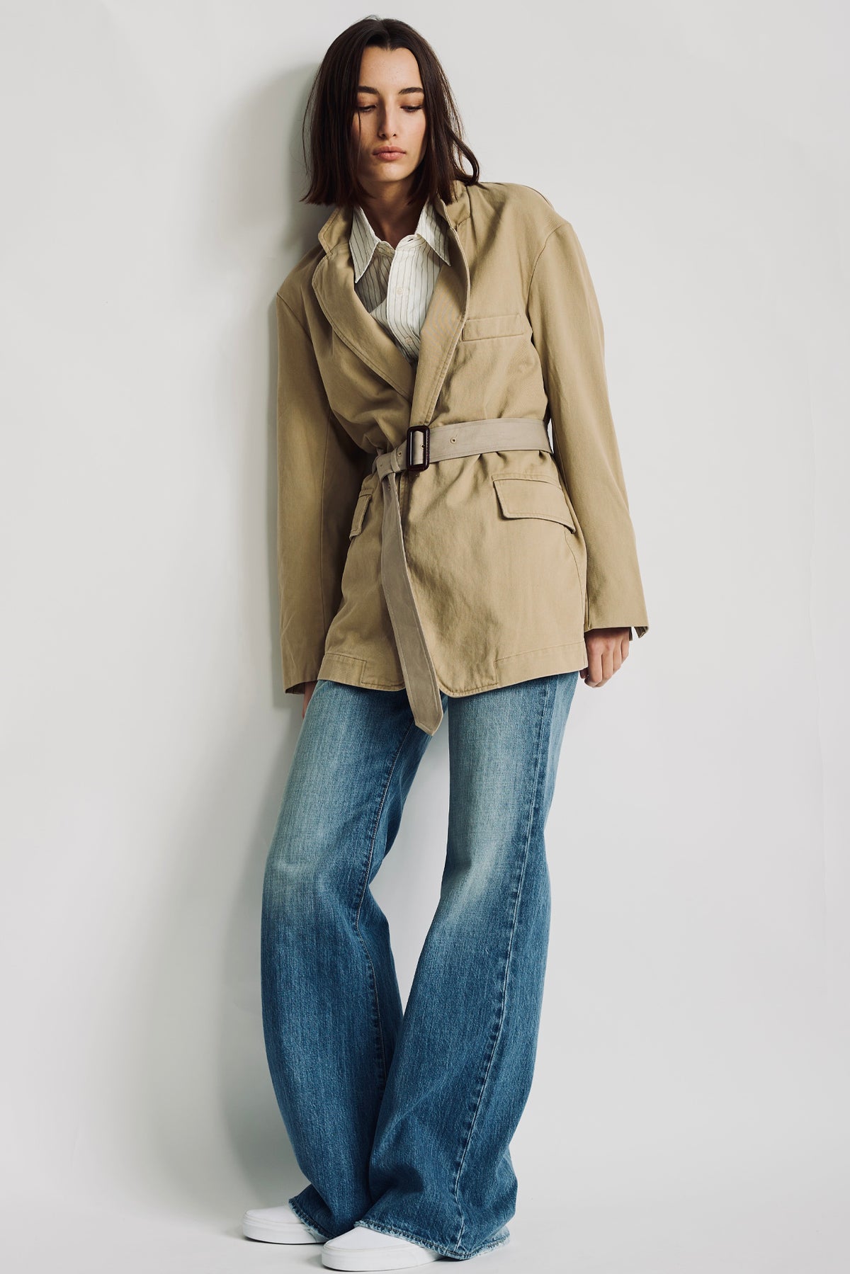 Cream button-front shirt with white double pinstripe visible under beige trench coat, worn with blue jeans.